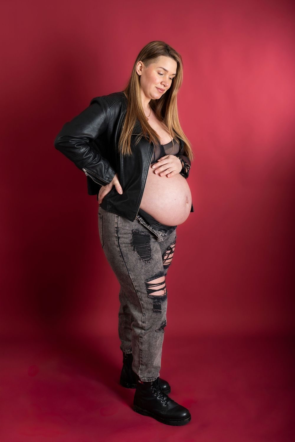 Pregnant woman in leather jacket and distressed jeans poses against red backdrop during maternity photoshoot.