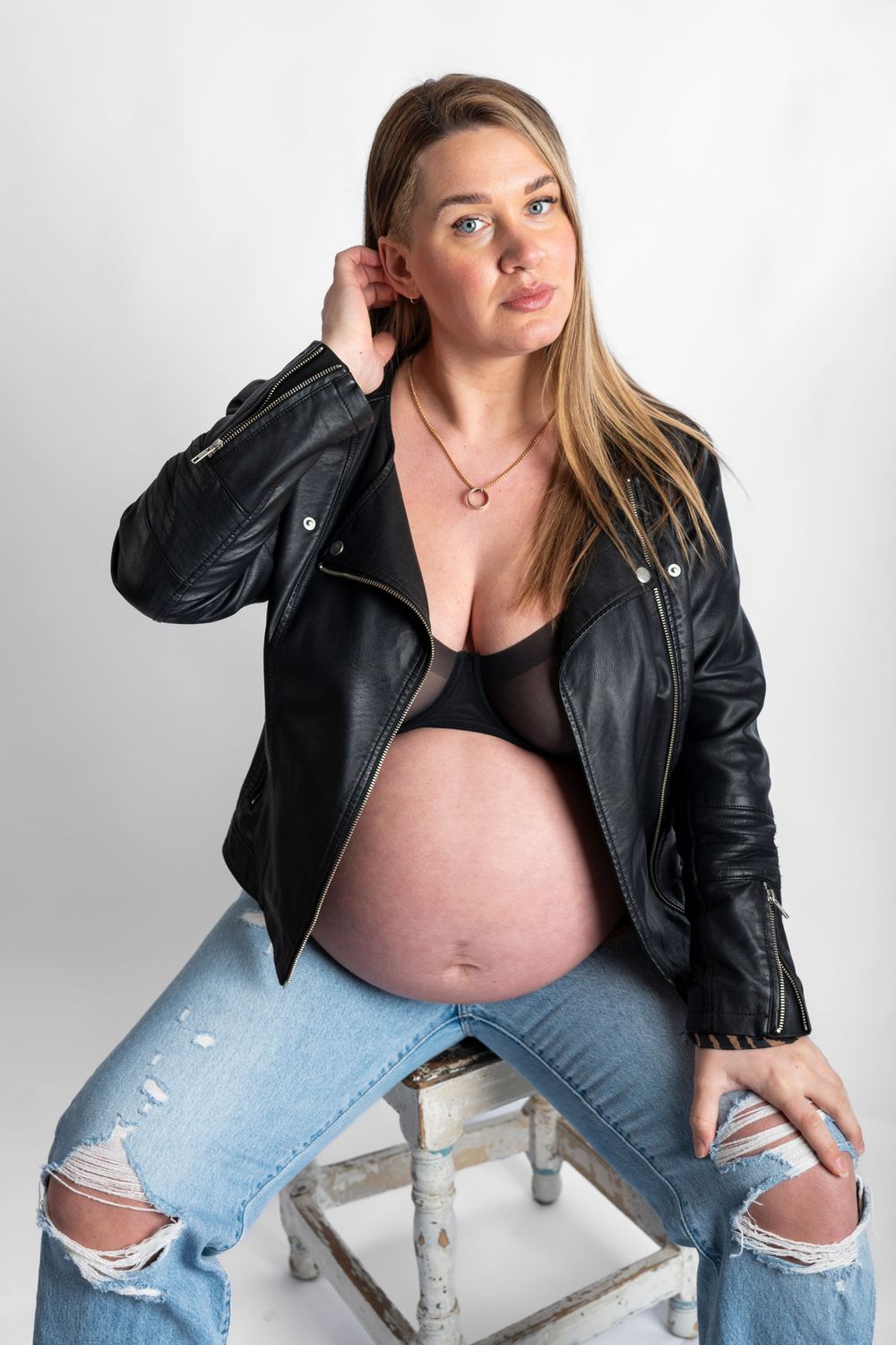 Expectant mother in leather jacket and ripped jeans poses casually against white backdrop in studio setting.