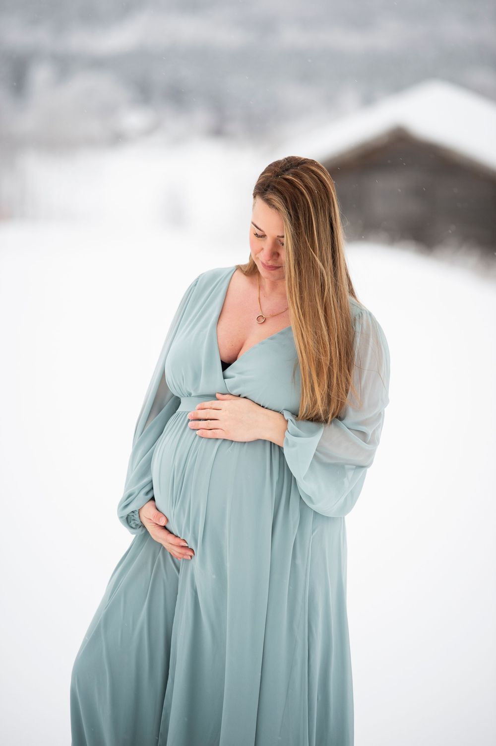 Expecting mother in flowing light blue dress poses in snowy winter landscape during maternity photoshoot.