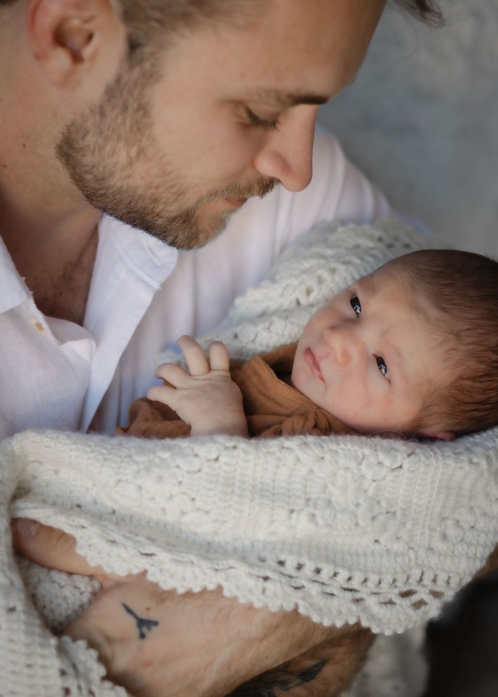 A tender moment showing parent and newborn baby wrapped in white knit blanket sharing an intimate close-up connection.