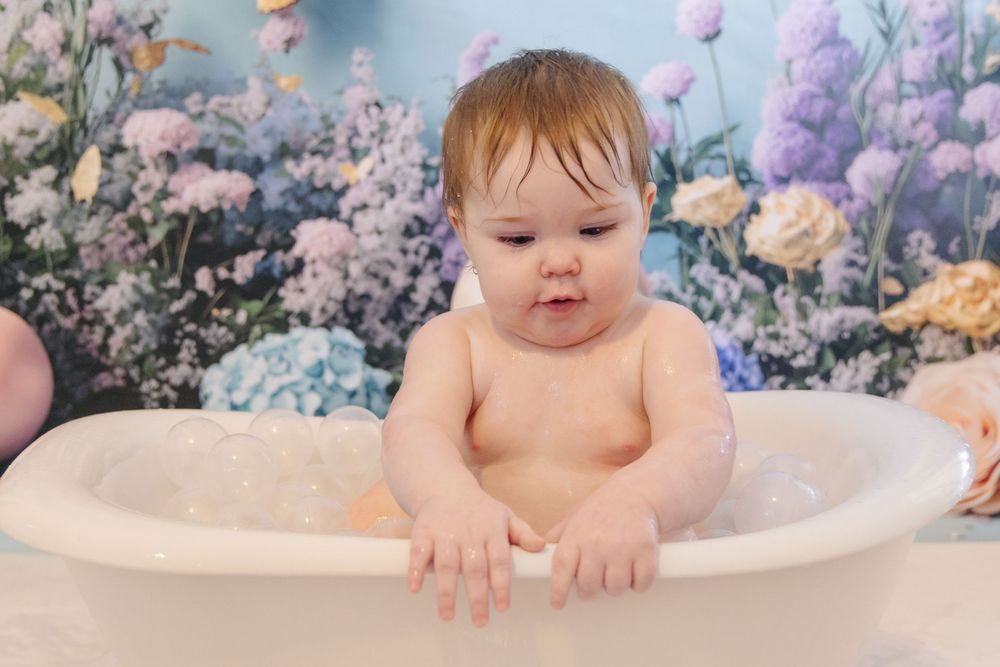 Baby playing in milky bath water surrounded by dreamy floral decorations in pastel colors.