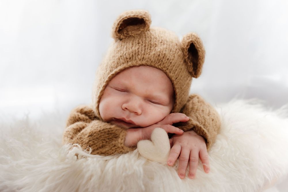 A tiny baby wearing a brown knit hat rests peacefully on soft white blankets.