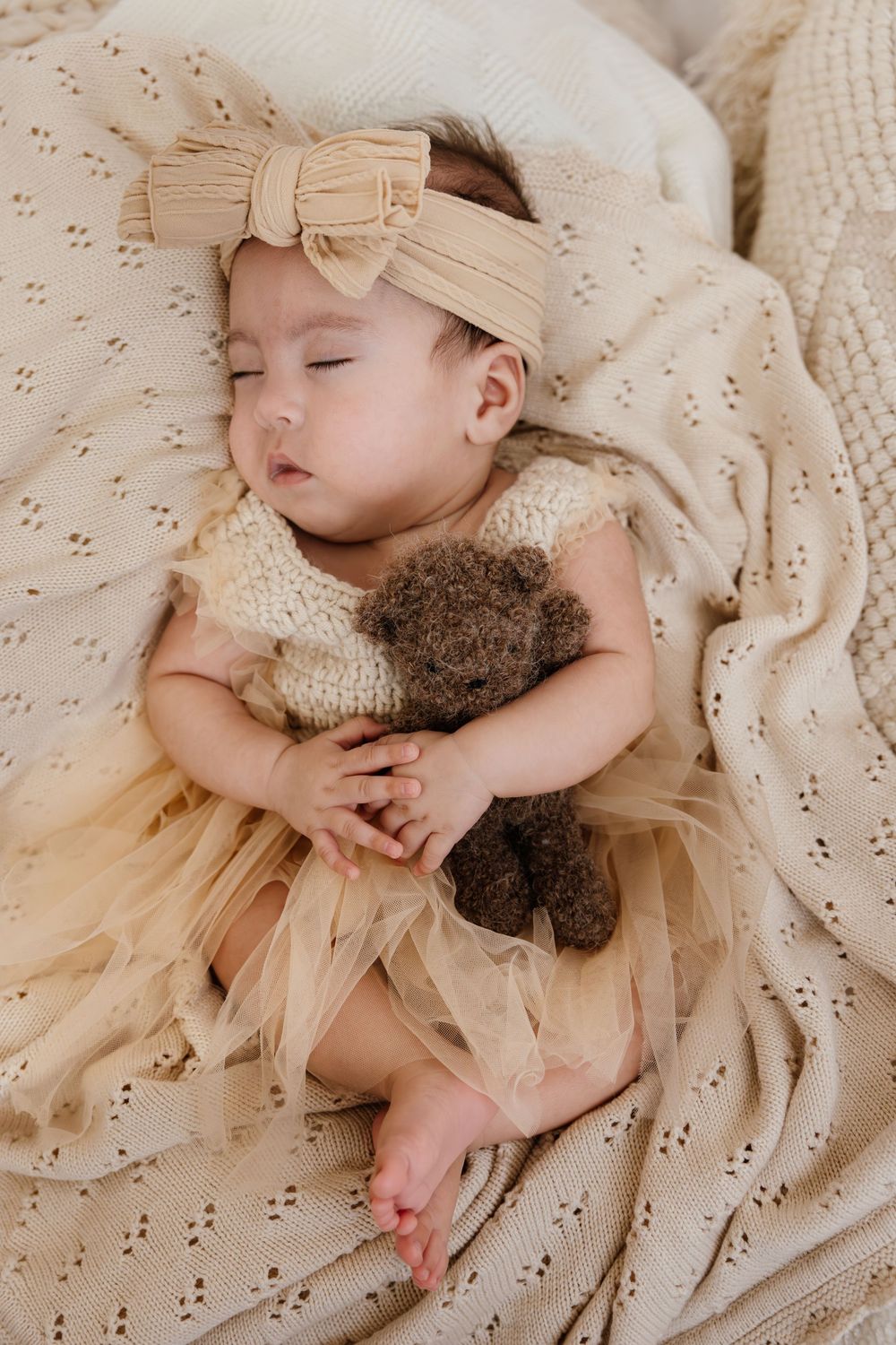 Baby sleeping peacefully on cream-colored eyelet fabric while cuddling a small brown teddy bear.