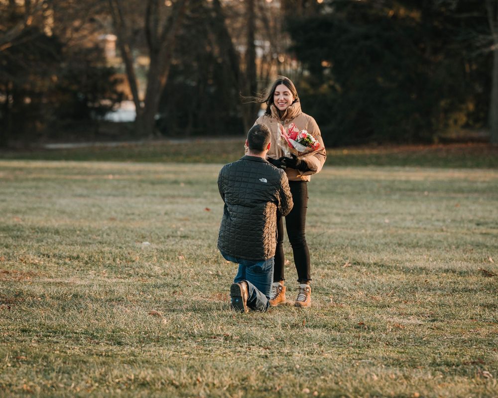 A romantic proposal scene unfolds on a grassy field during golden hour as two people share a special moment.