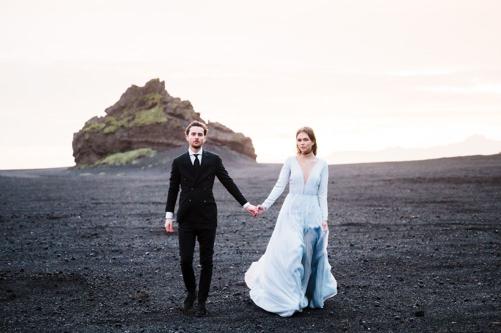 A couple in wedding attire walks hand in hand on a black sand beach with a dramatic rocky outcrop in Iceland.