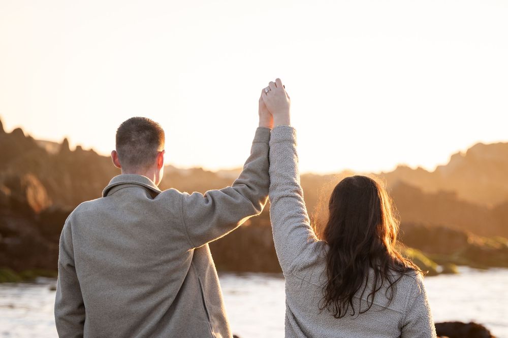 A couple stands together on a beach at sunset with arms raised in celebration.