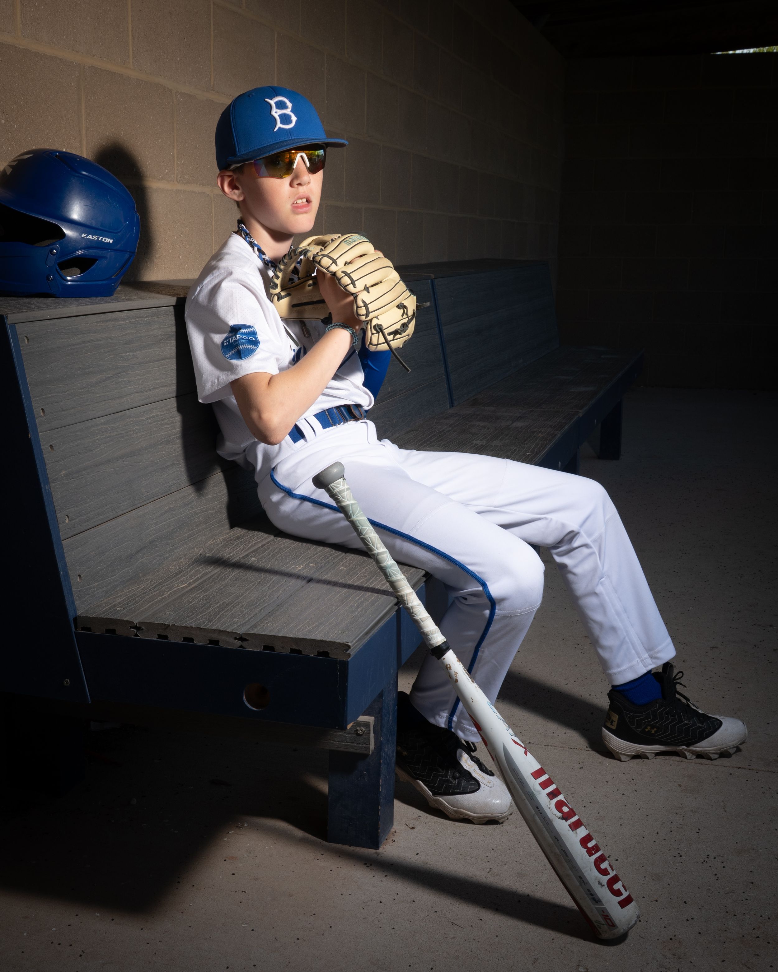 A baseball player sits in a dugout wearing a blue cap and white uniform in dramatic lighting.