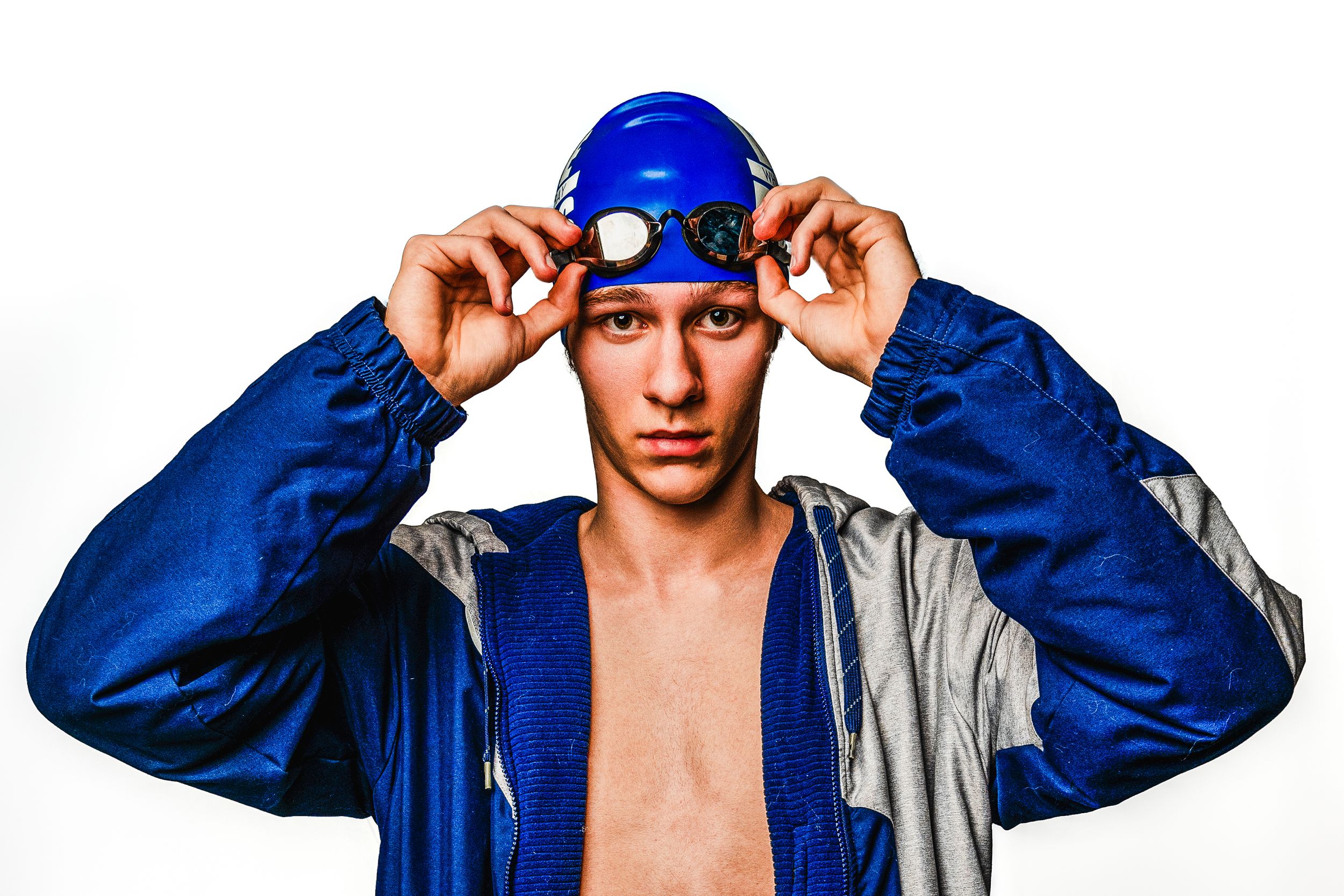 Athletic swimmer in blue swim cap and jacket adjusts gear against white background.