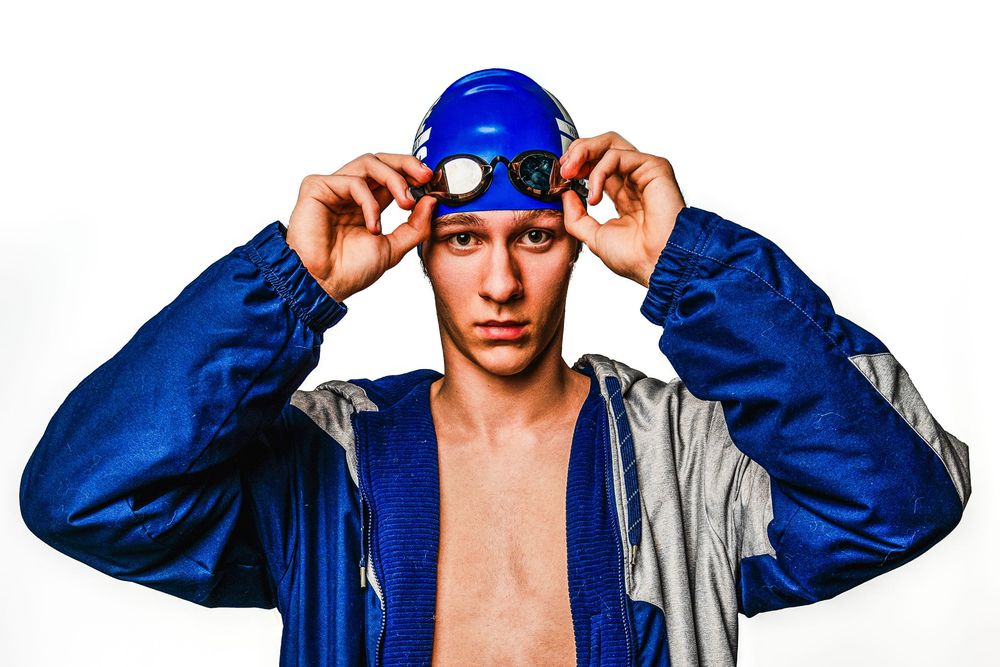 Athletic swimmer in blue swim cap and jacket adjusts gear against white background.