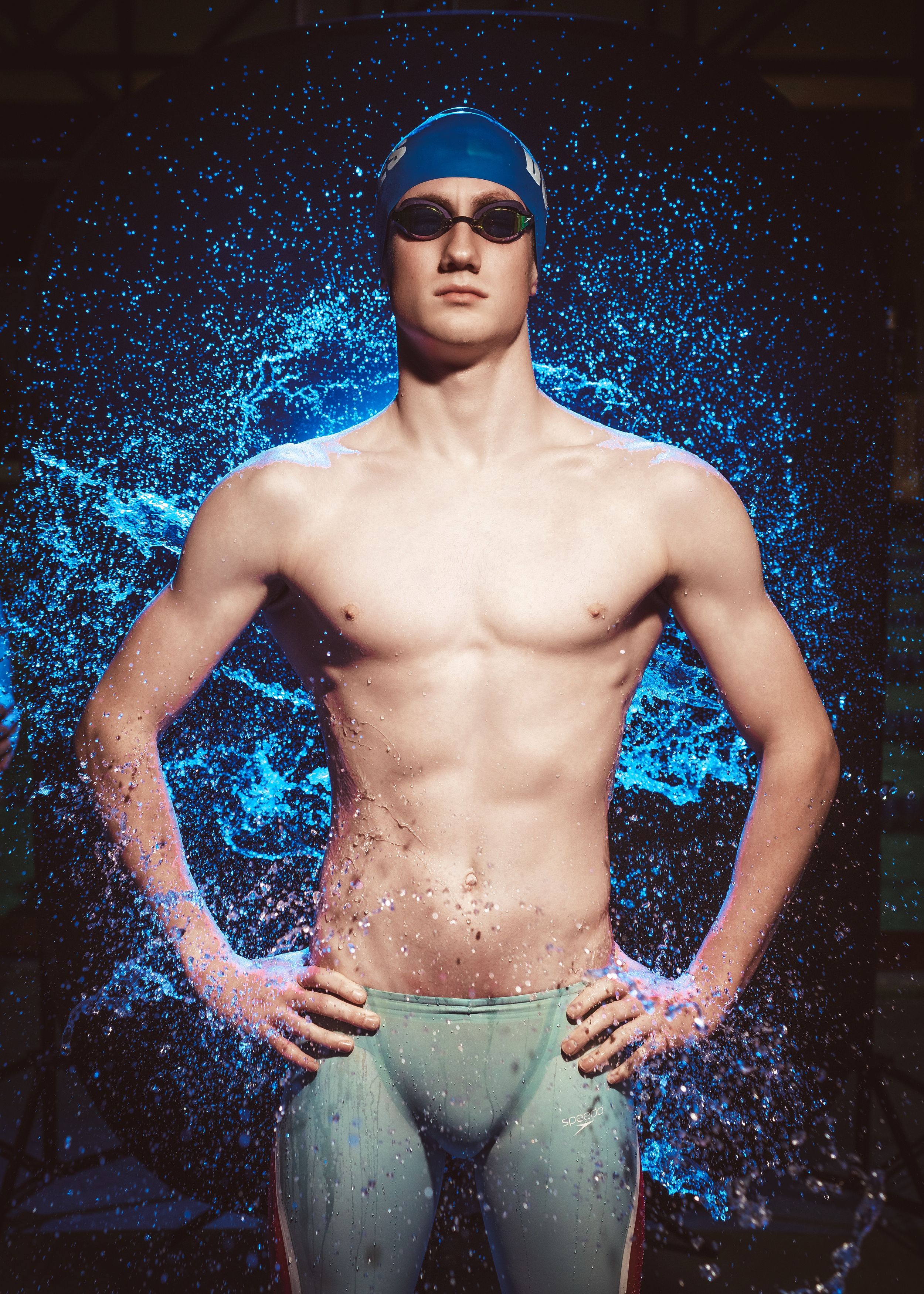 Athletic swimmer in blue swim cap and goggles stands against dark background with splashing water effects in bright blue.