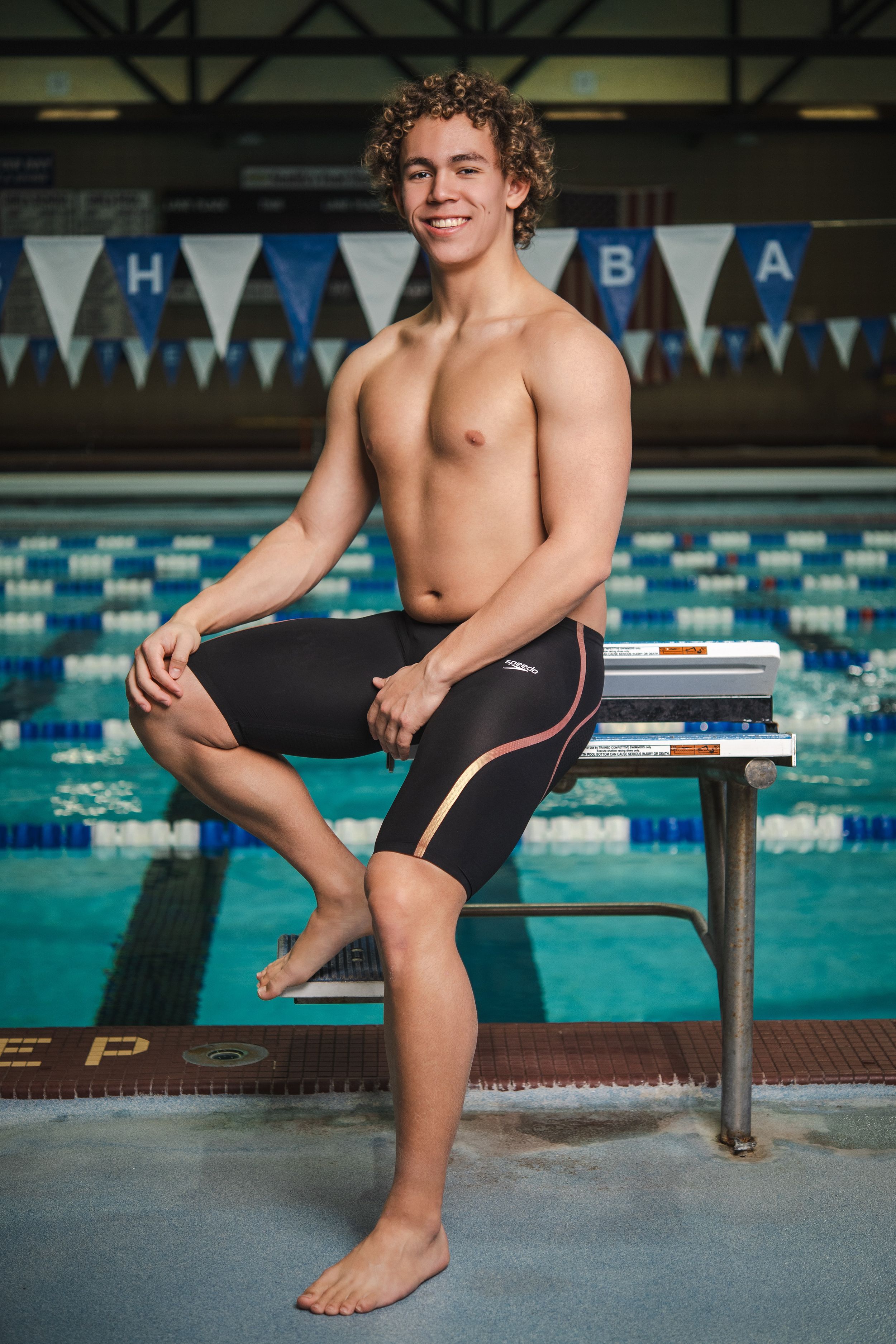 Athletic swimmer in black jammers sits on diving block at indoor pool with blue lane markers and competition banners.