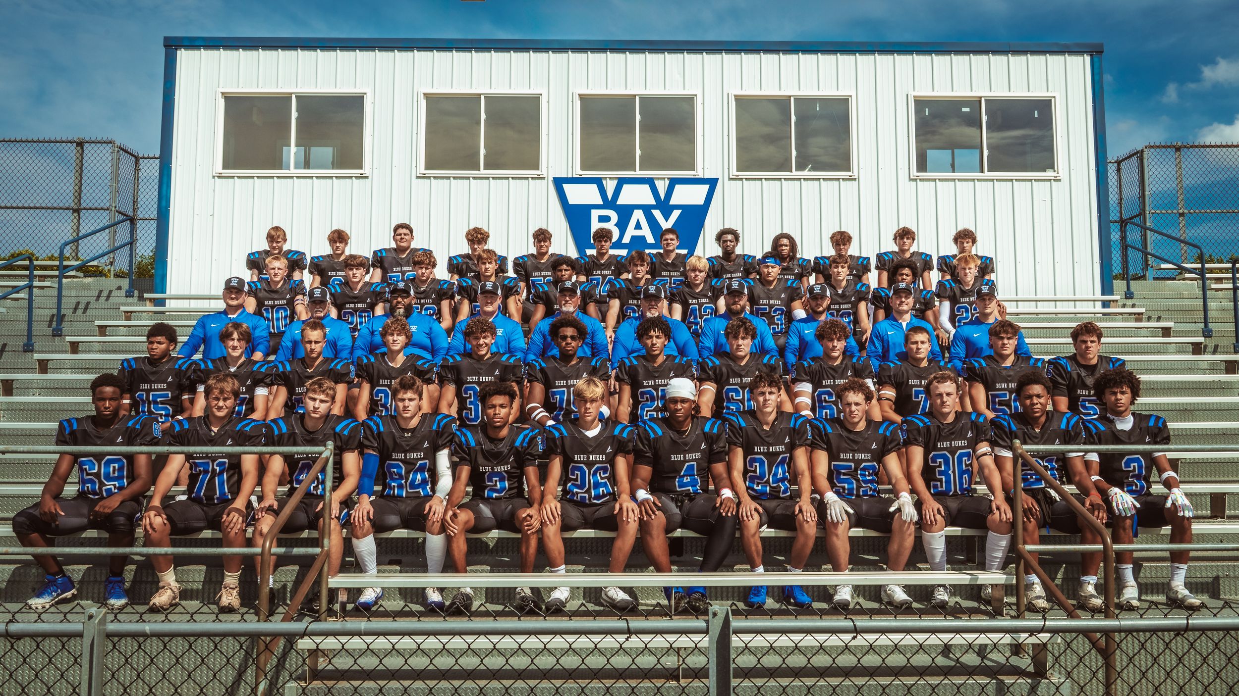 Large group of athletes in blue uniforms sit together on bleachers for team photo session.