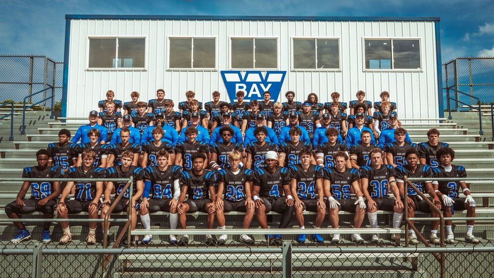 Large group of athletes in blue uniforms sit together on bleachers for team photo session.