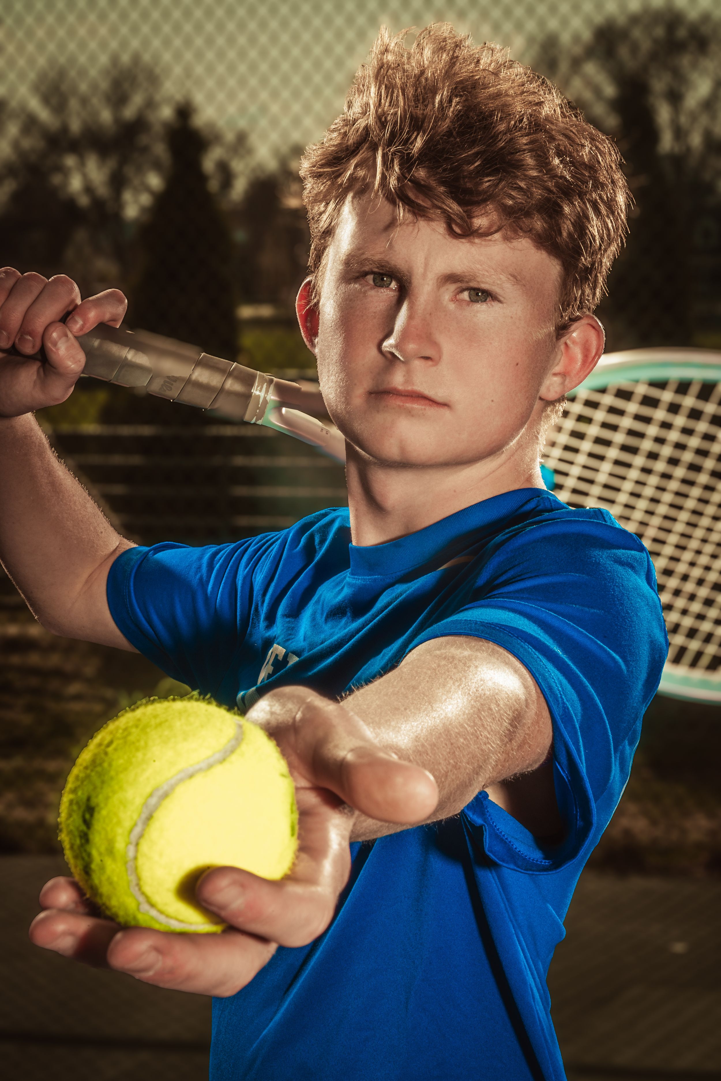 Young tennis player in blue shirt poses with racquet and tennis ball during golden hour outdoor photo shoot.