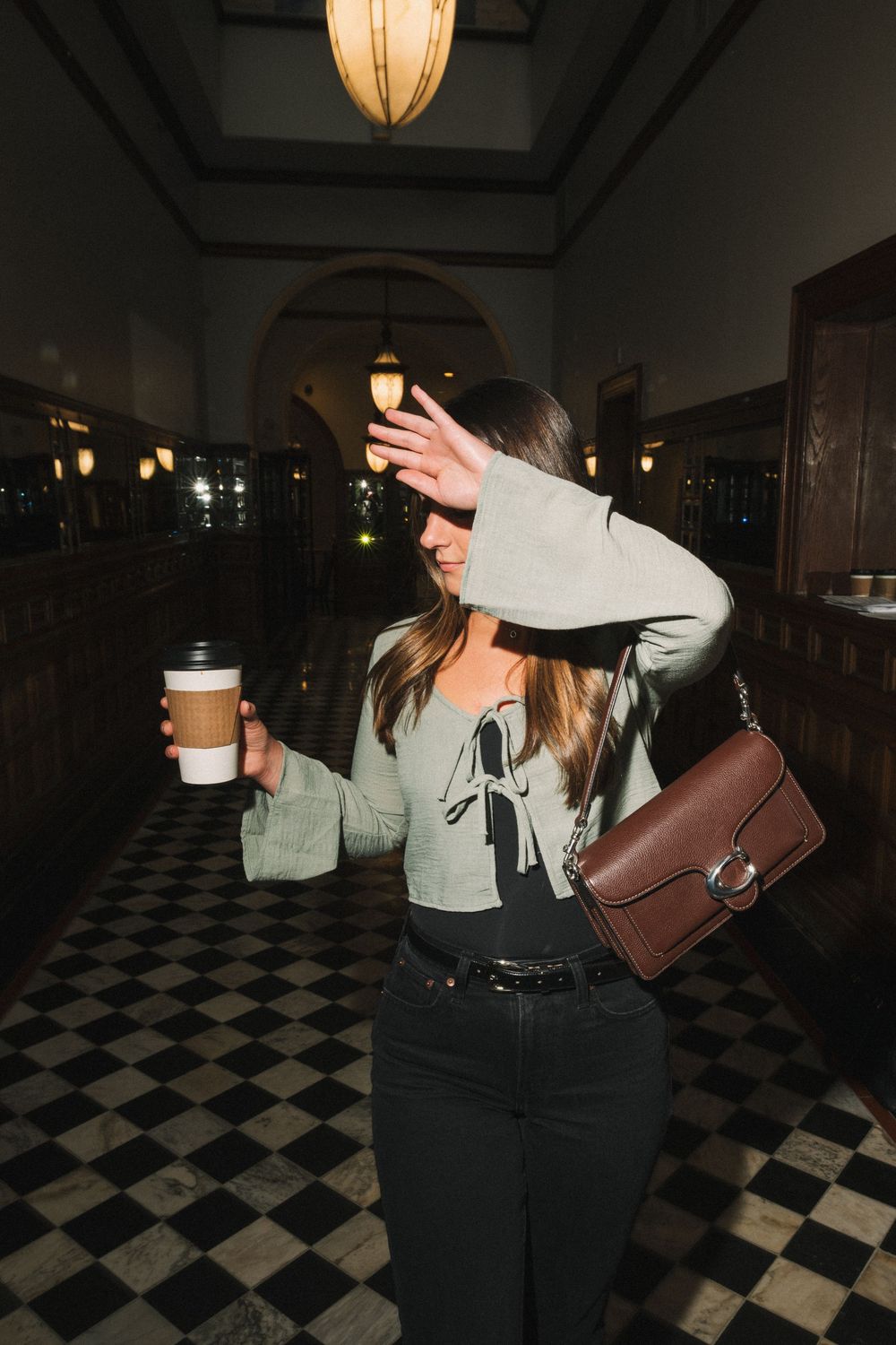 Person walking on checkered floor with coffee cup and brown leather bag.