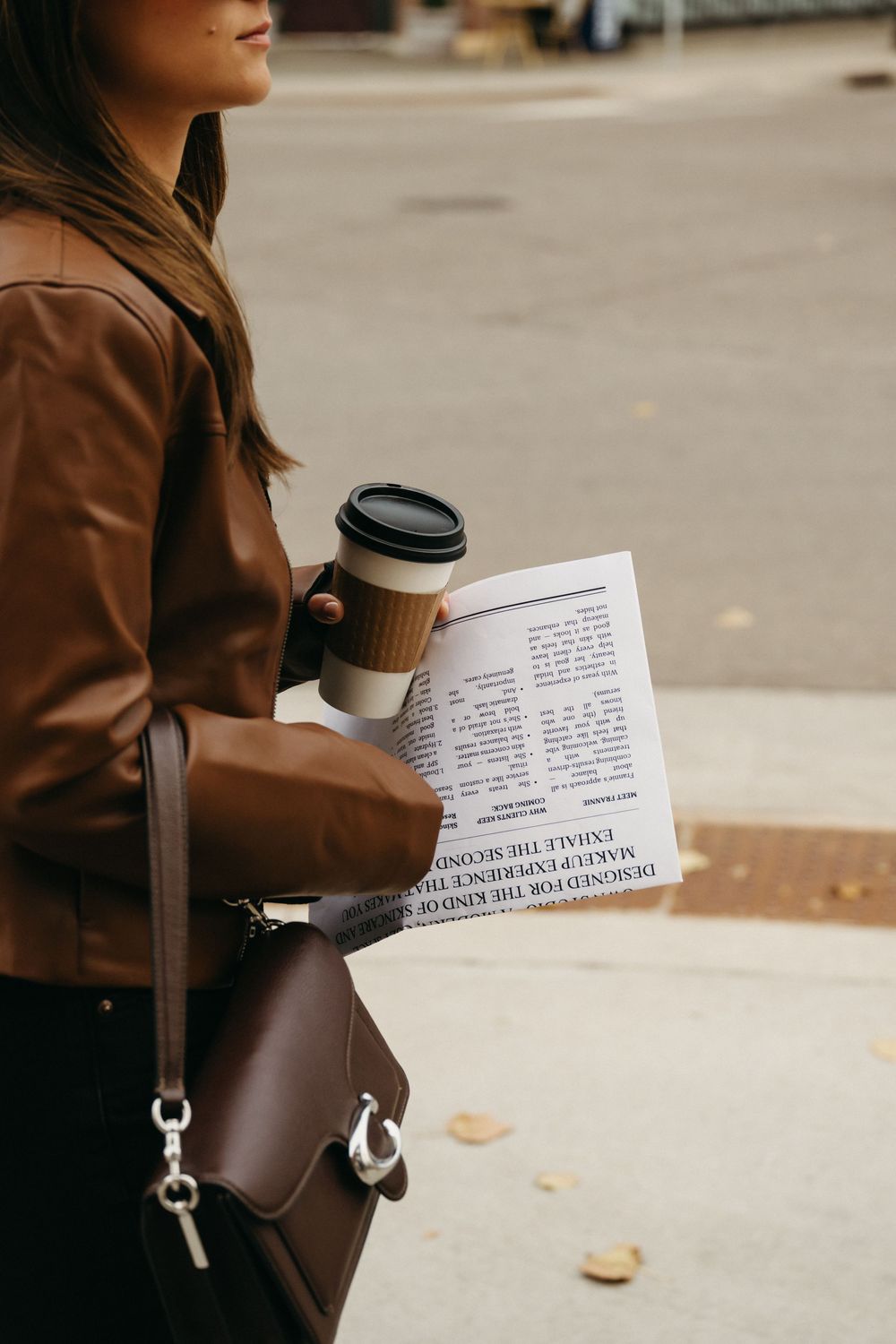 Person in brown jacket holding coffee and papers while walking past brick buildings.