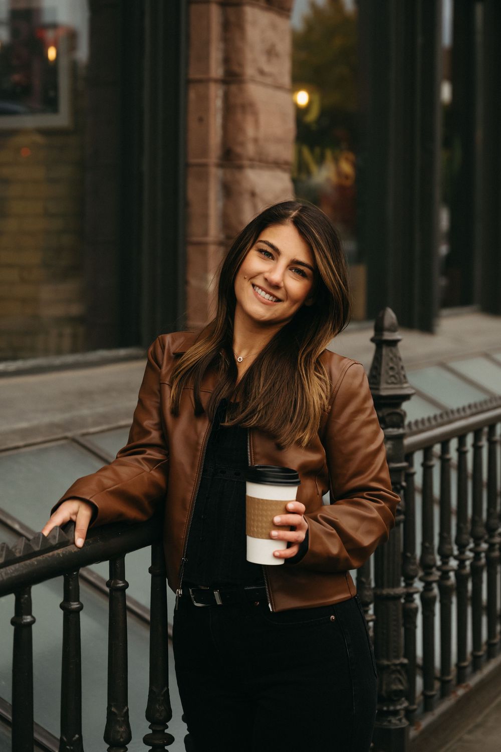 Person in brown leather jacket holding coffee cup while leaning on outdoor railing.