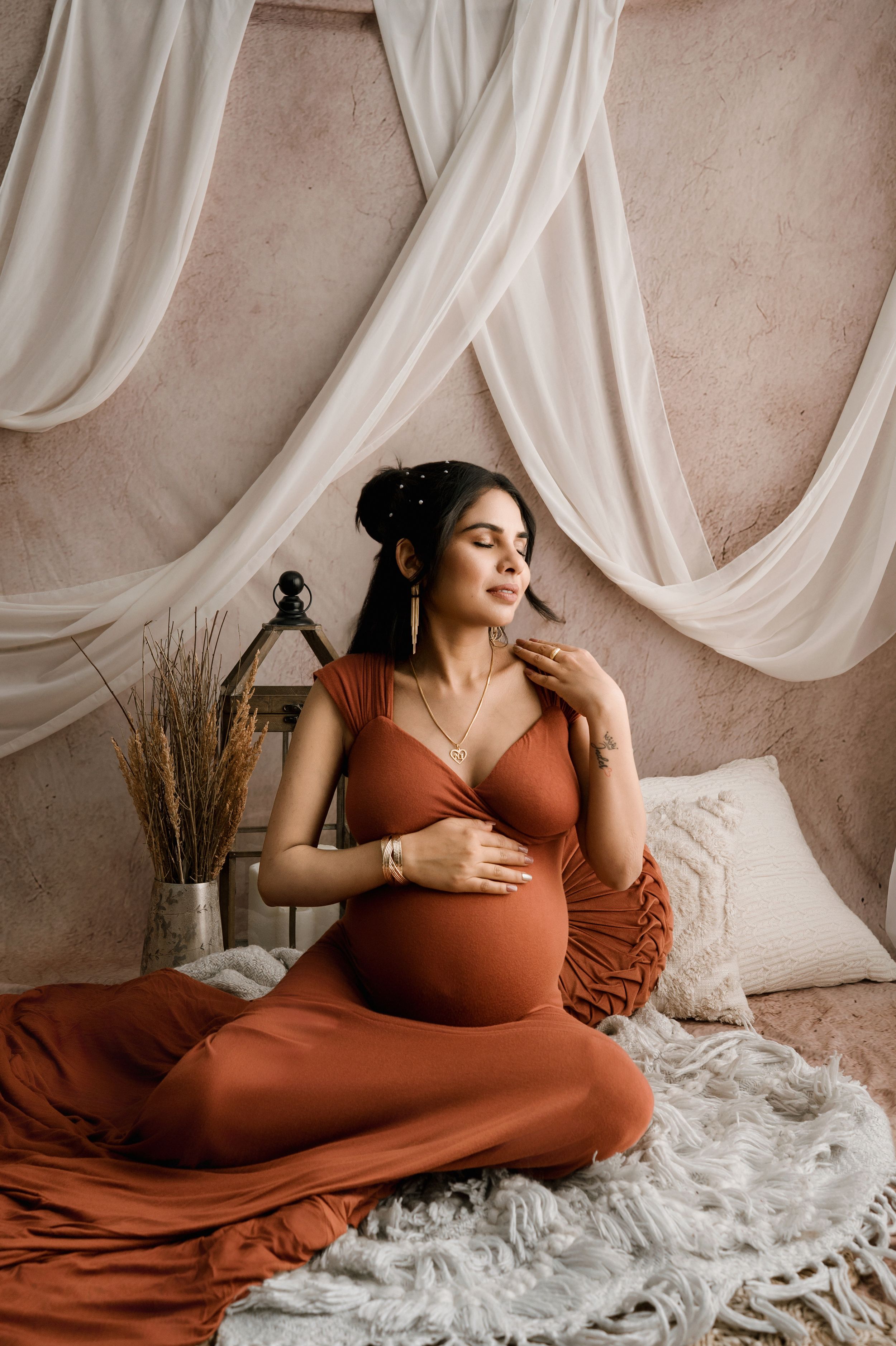 A person in a rust-colored dress reclines on white bedding with flowing fabric draped in the background.