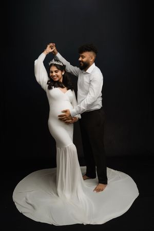 A couple in wedding attire dance together against a dark background during a romantic maternity photo session.