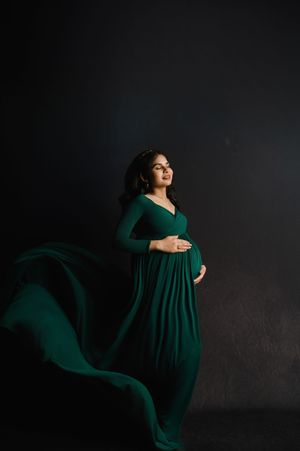 A flowing emerald green maternity gown creates an elegant silhouette against a dark background in a dramatic portrait.