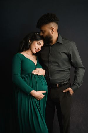 Intimate maternity photo against dark backdrop features couple in green dress and grey shirt embracing tenderly.