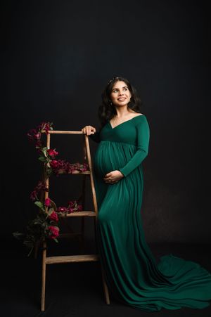A maternity photo series in a dark studio features a flowing emerald green dress and wooden chair with floral accents.