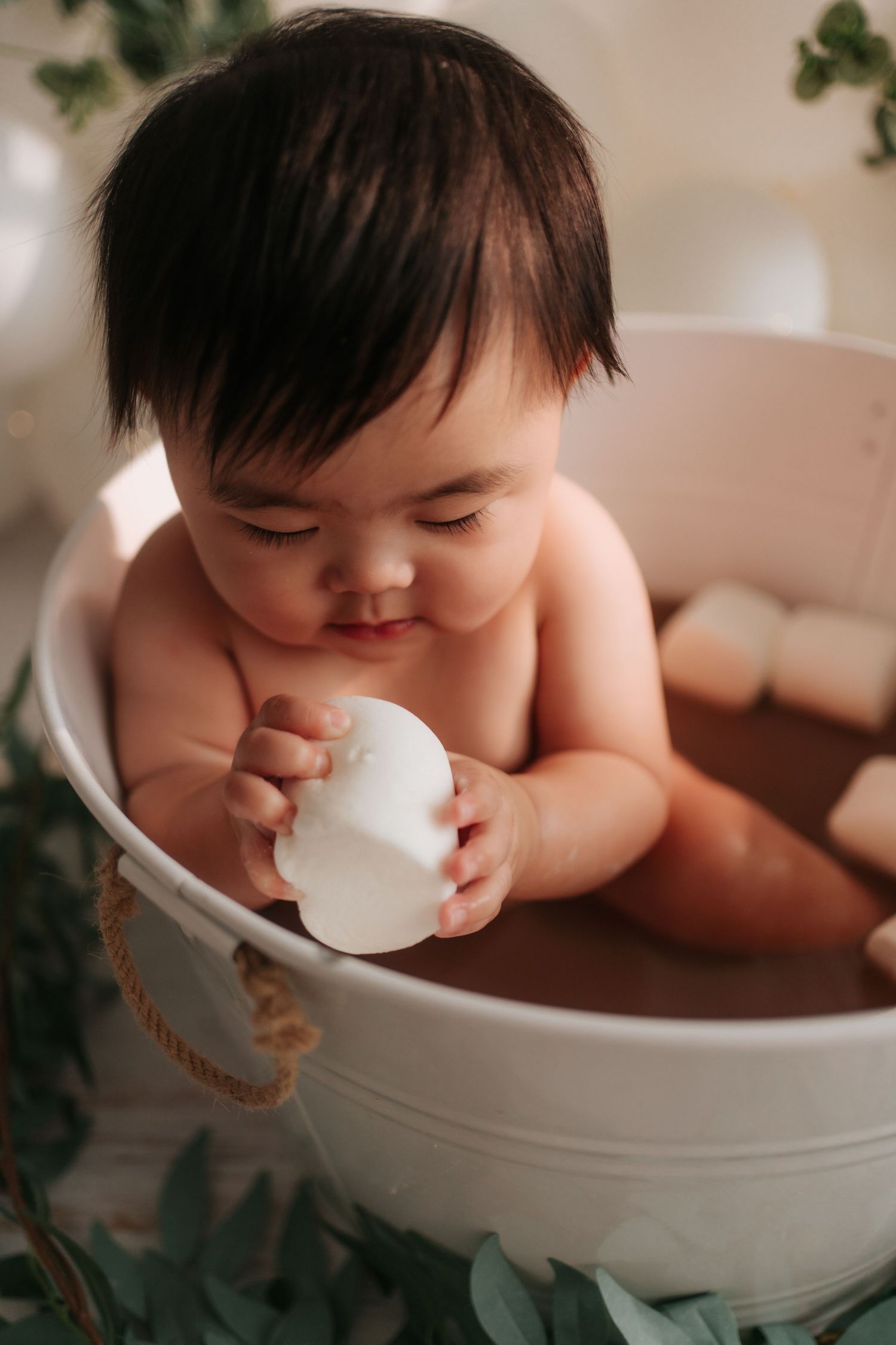 Baby takes a bath in white ceramic tub while playfully drinking from white cup.