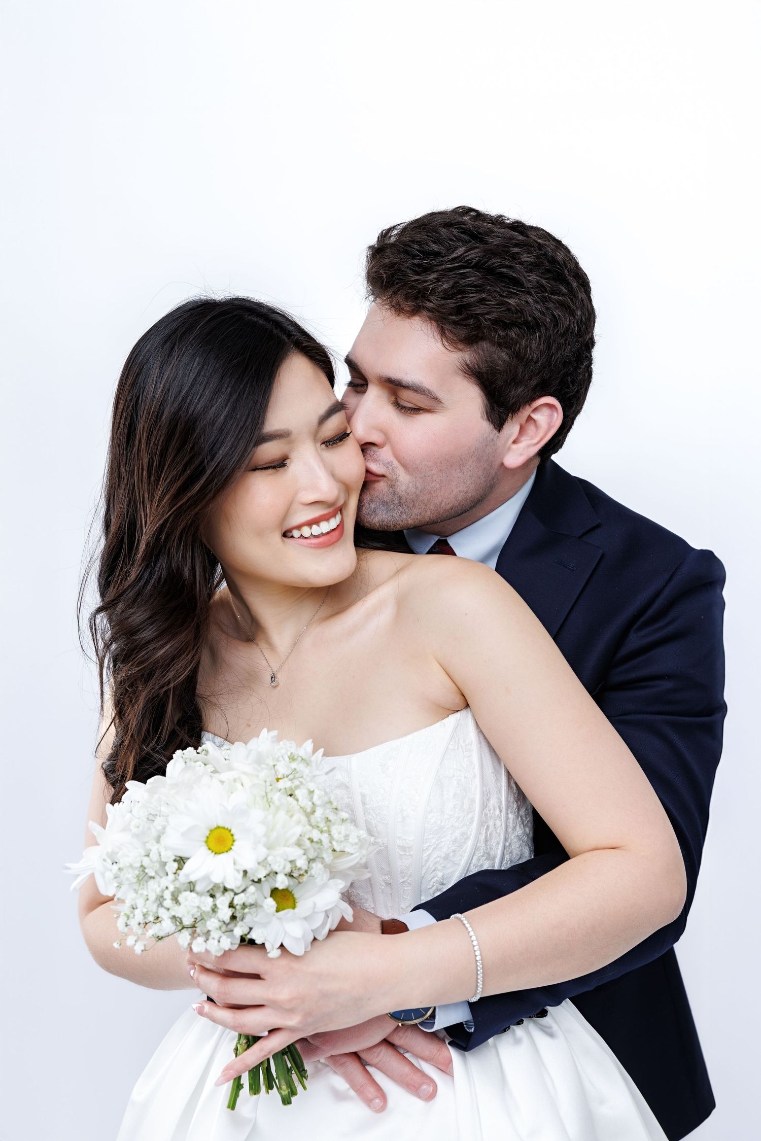 A couple in wedding attire poses with their brown dog wearing a bow tie on a white backdrop for formal portraits.