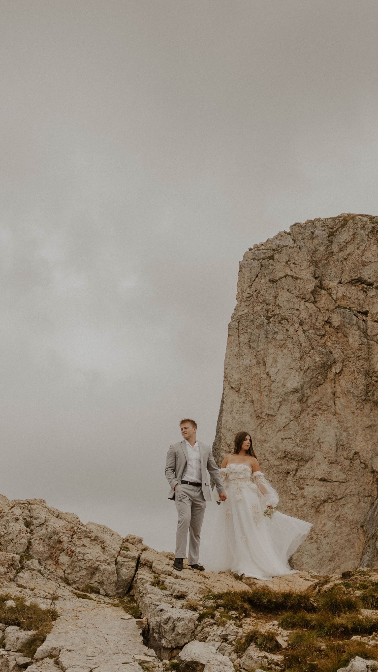 A couple in wedding attire stands atop a dramatic rocky cliff under an overcast sky in a moody mountainous landscape.