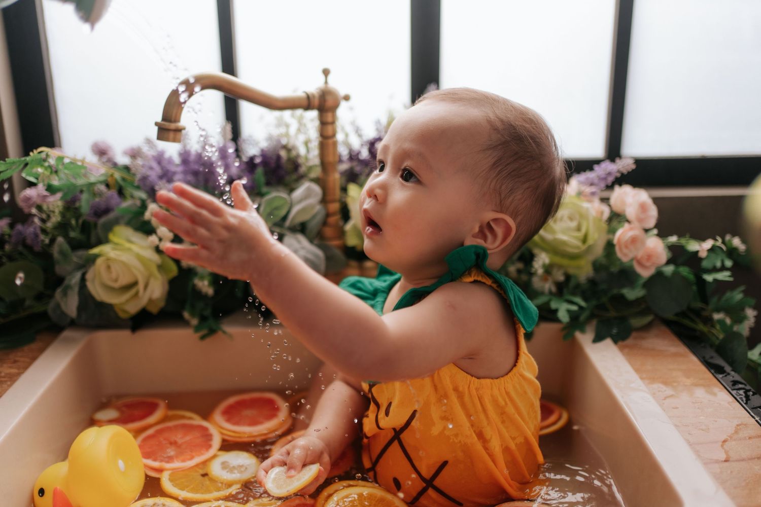 A toddler in an orange shirt reaches for food and drinks at a dining table.