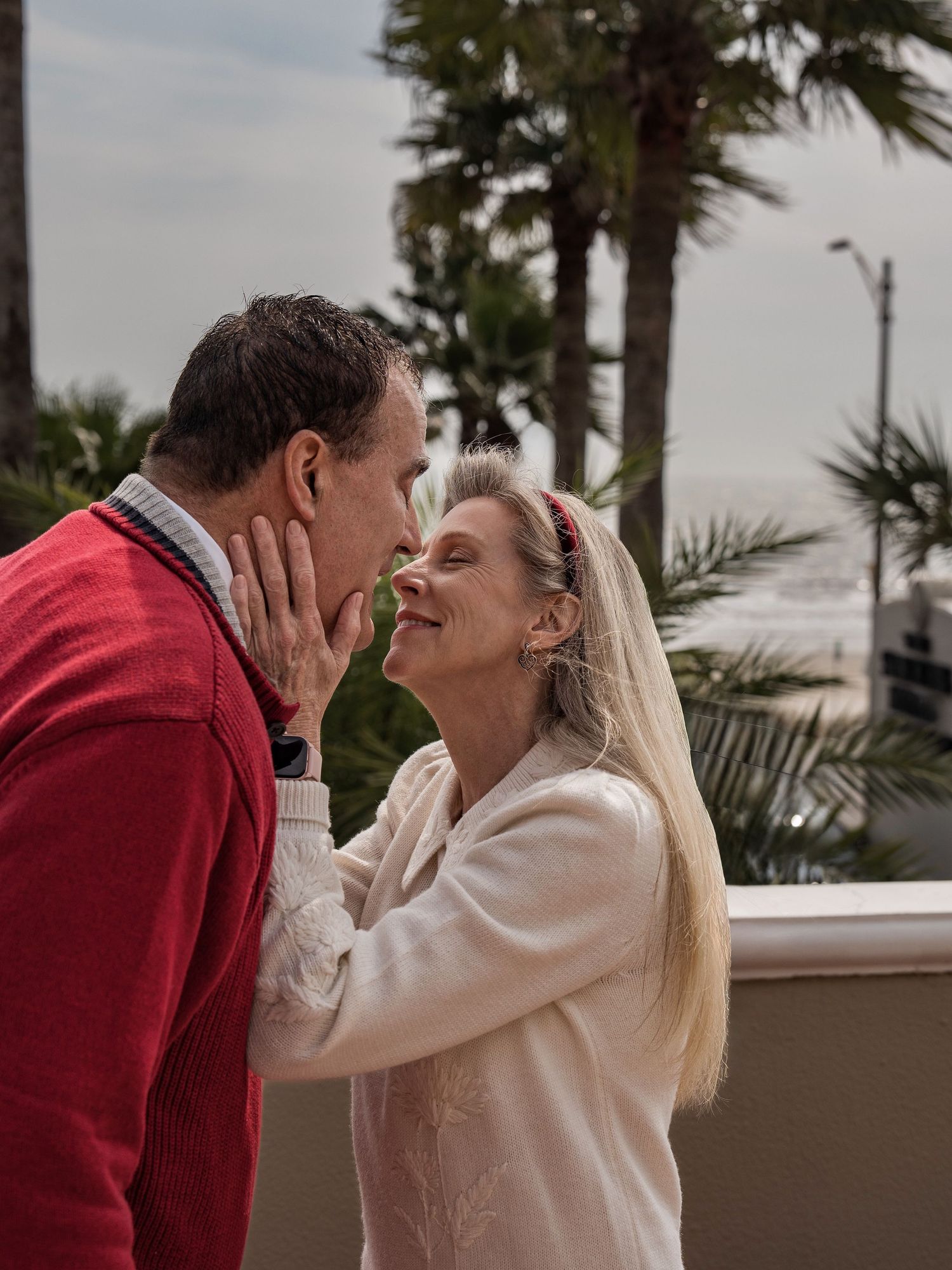 A romantic couple shares intimate kisses on a balcony with palm trees in the background.