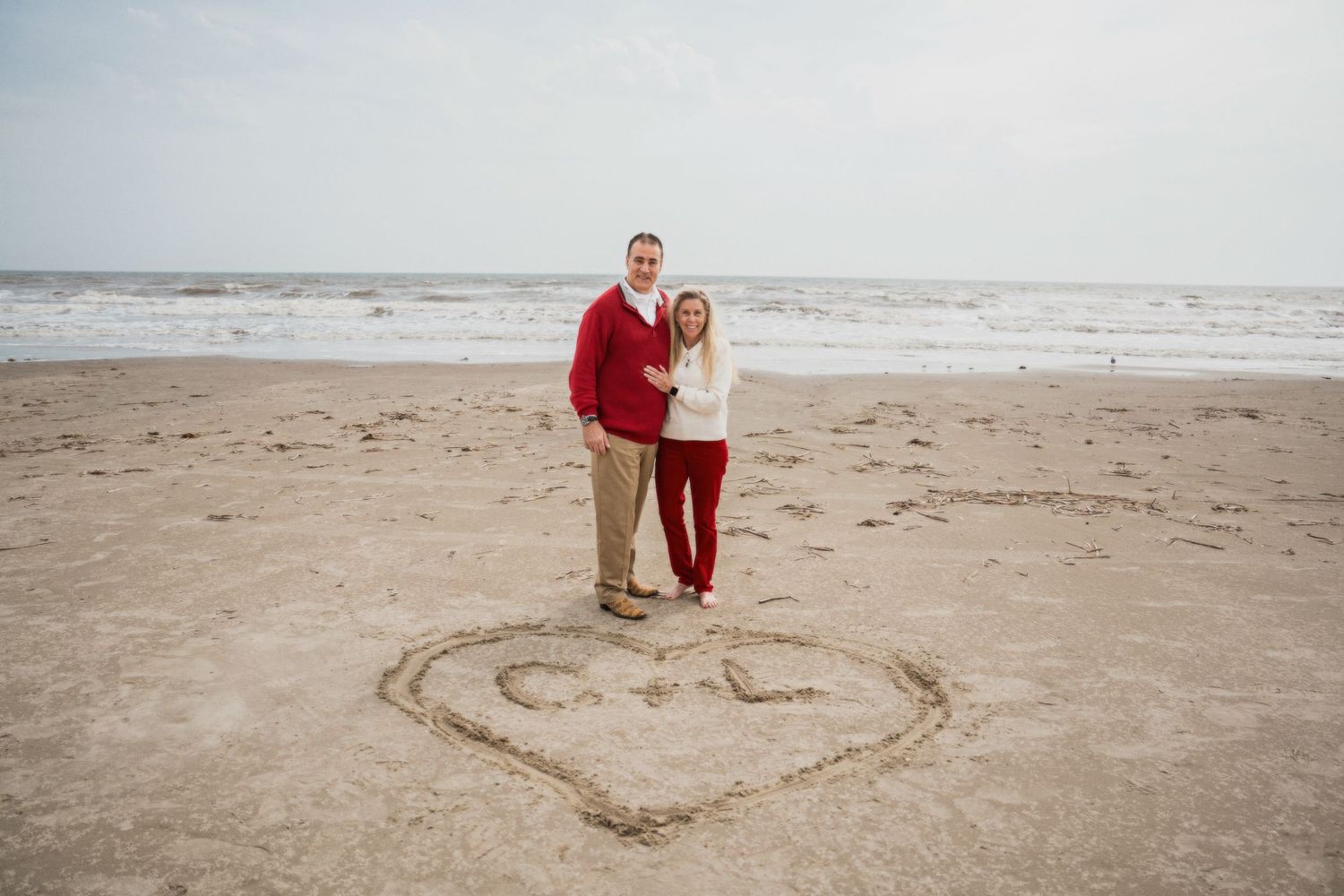 Two people draw a heart shape in the sand on a cloudy beach during a romantic photoshoot.