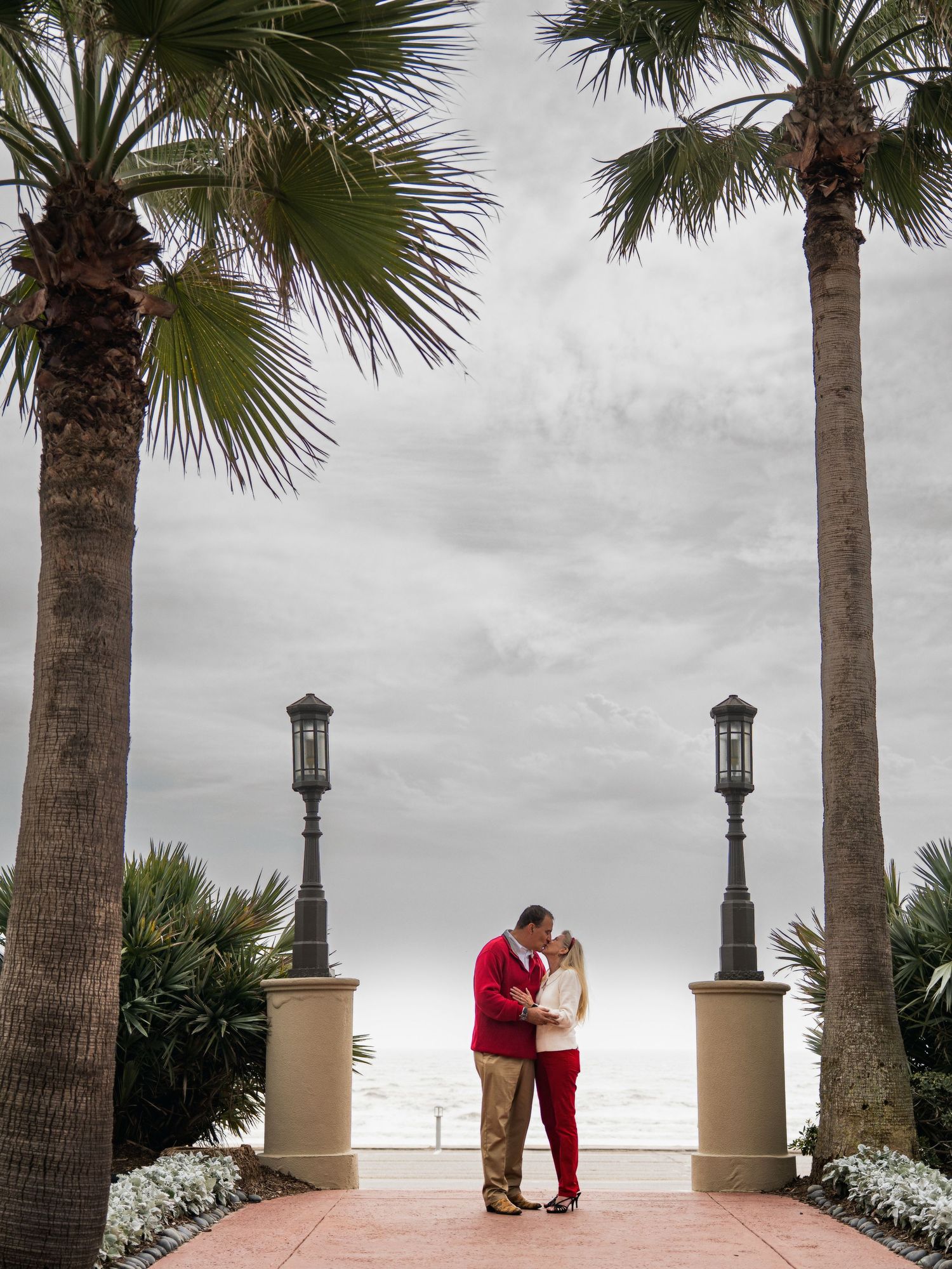 A romantic scene unfolds beneath palm trees with a cloudy coastal backdrop.