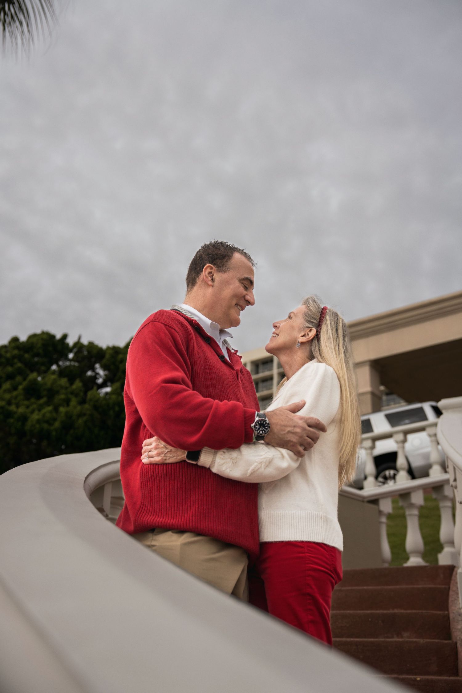 A romantic couple embraces on outdoor steps in coordinating red and white sweaters on an overcast day.