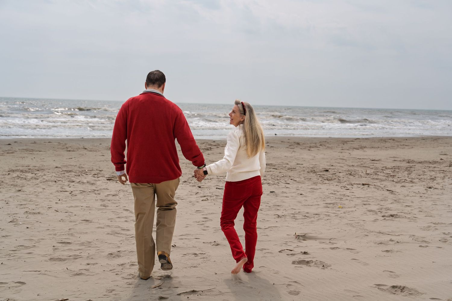 A couple takes a romantic stroll along a windy beach while wearing coordinating red and cream outfits.