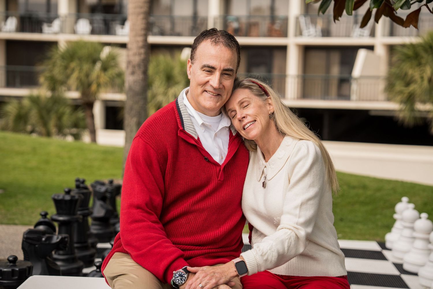 Couple in red and white sweaters sitting together on outdoor chess board near beach condos.