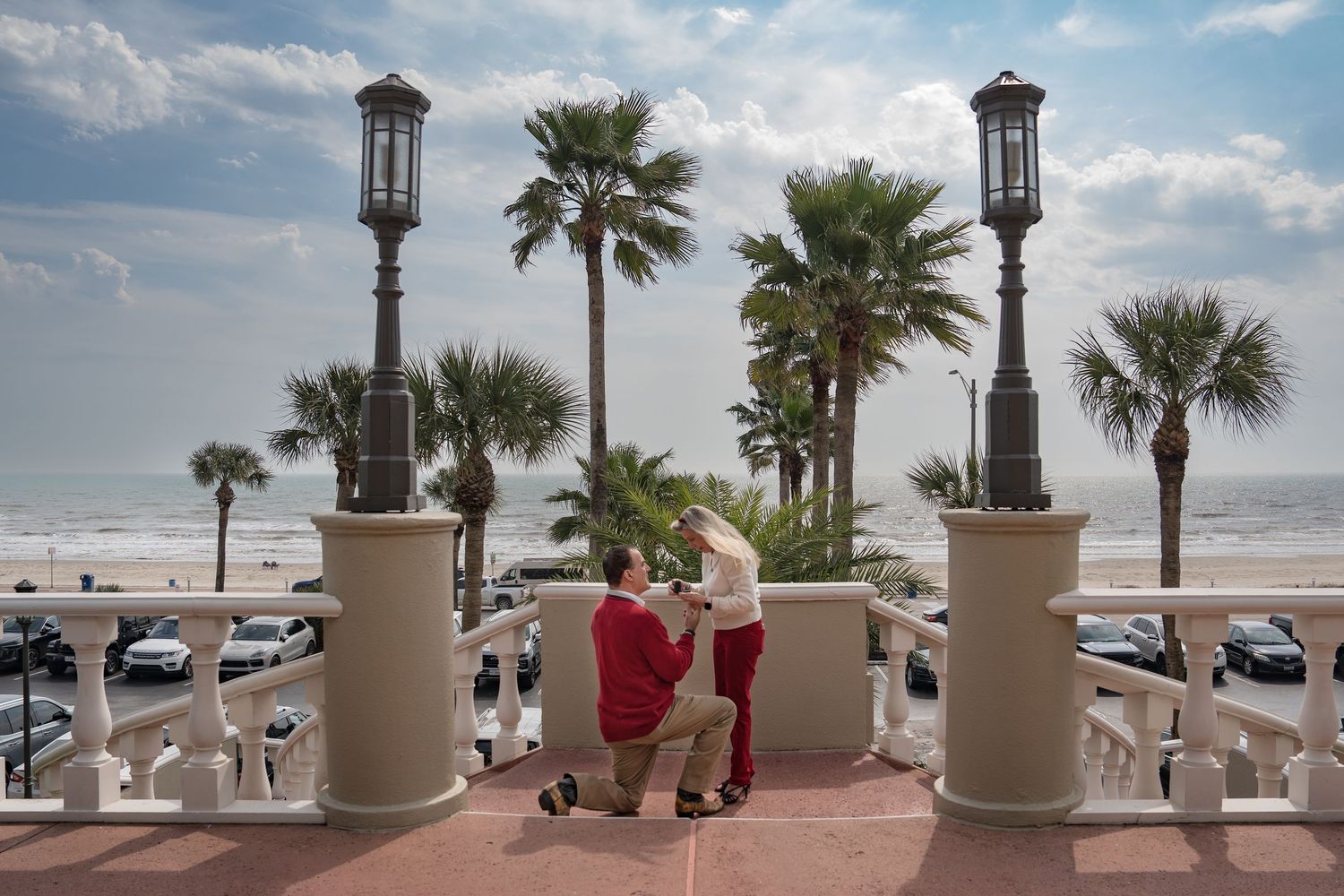 Couple stands between ornate lamp posts on a beachfront promenade overlooking palm trees and parked cars.