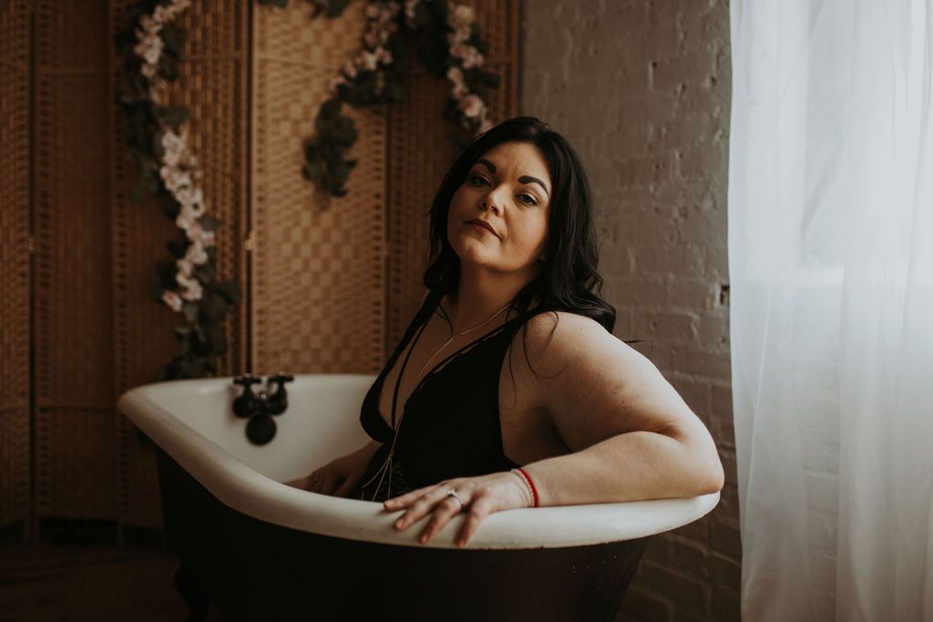 A woman poses elegantly in a bathtub with white curtains and floral decorations creating a dreamy atmosphere.
