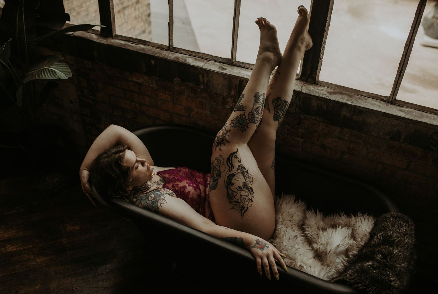 A curvy woman with tattoos wears a burgundy bodysuit while relaxing in a vintage clawfoot bathtub with her feet propped up