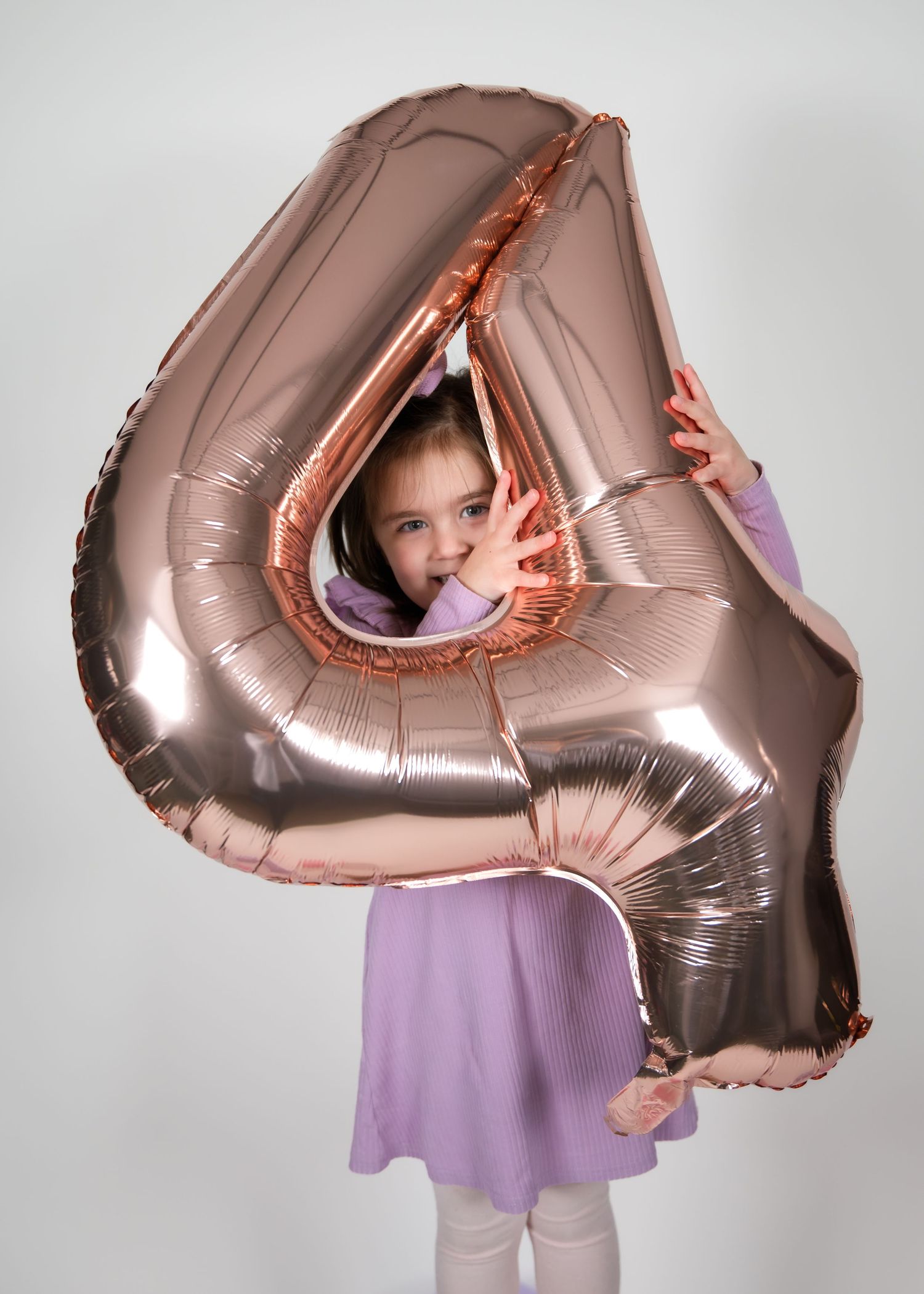 Four sequential photos show someone playfully peeking through a large rose gold heart-shaped balloon against a light background.