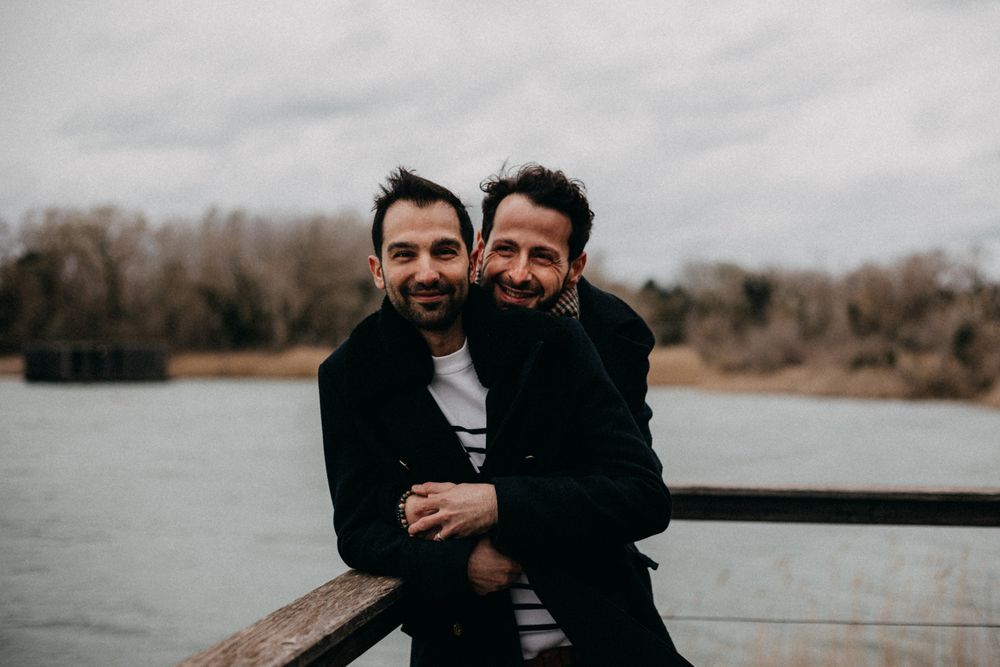 Two people laugh and embrace while standing on a wooden pier with a lake and bare trees in the background.