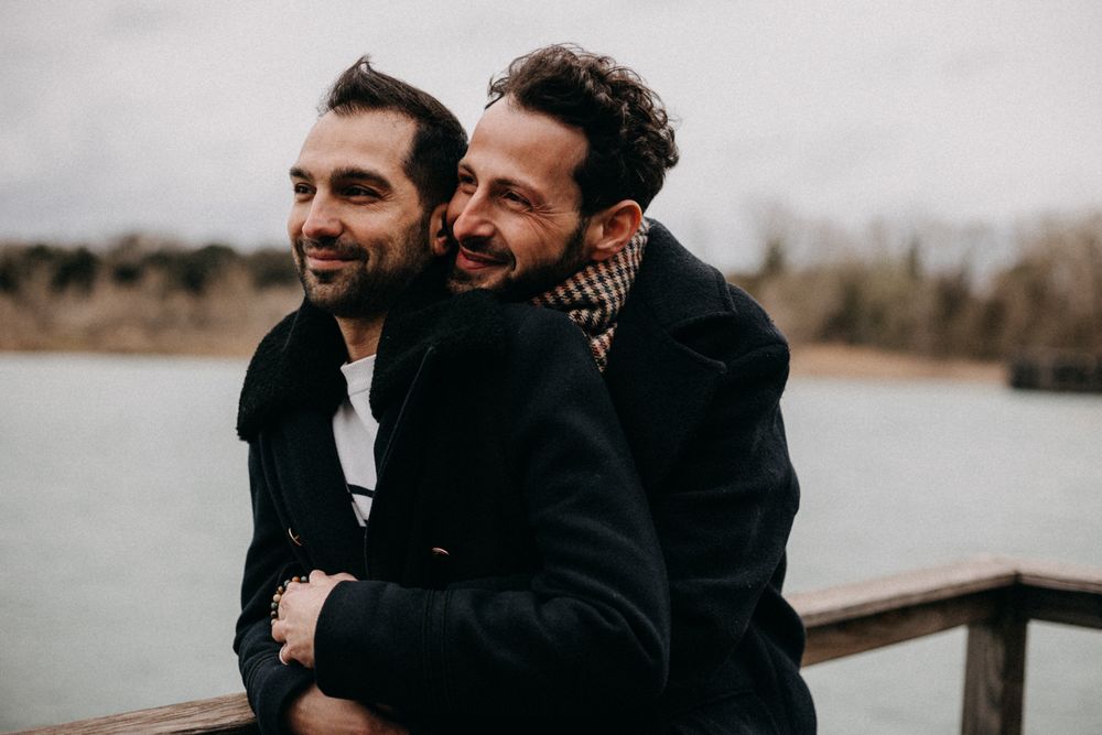 Two people share joyful laughter while sitting close together on a lakeside dock in winter.
