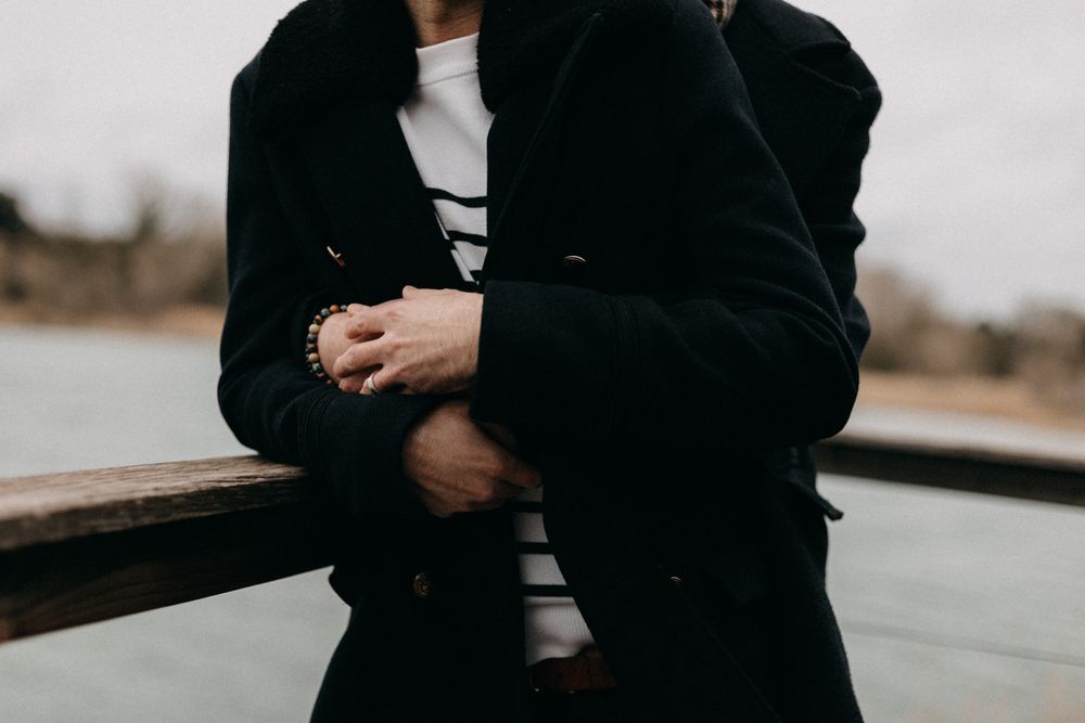 Person in black coat and white shirt stands with hands crossed against blurred outdoor background.