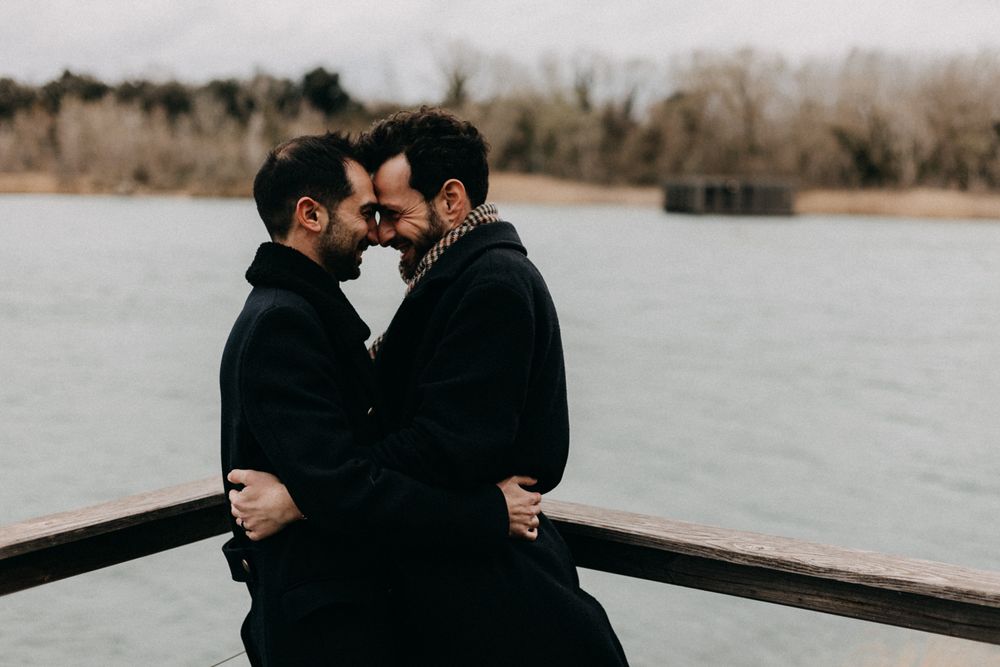 Two people in black coats share an intimate moment on a dock by a misty lake.