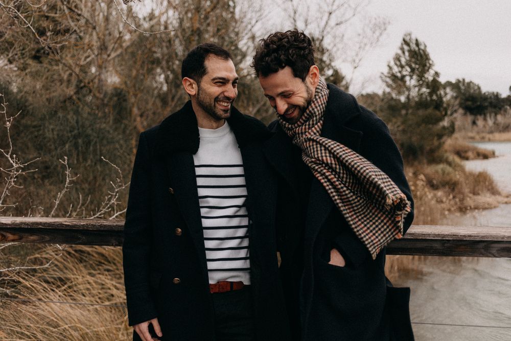 Two people share a tender moment by a waterfront on an overcast day wearing dark winter coats.