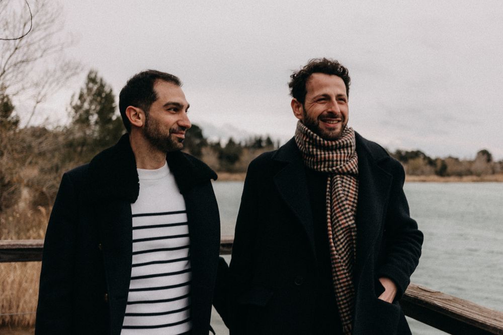 Two people in dark coats and scarves embrace while walking along a lakeside path on a cloudy day.