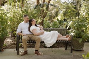 A couple relaxes together on a garden bench surrounded by lush greenery and white flowers on a sunny afternoon.