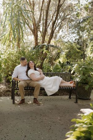A couple relaxes on a garden bench surrounded by lush tropical plants and palm trees in a romantic outdoor setting.