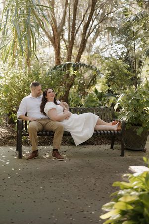 A couple relaxes together on a garden bench surrounded by lush greenery and dappled sunlight.