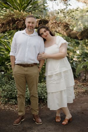 A couple poses together in a garden setting wearing coordinated white and khaki outfits among tropical foliage.